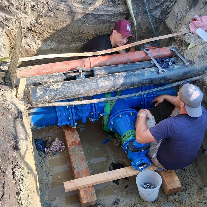 Two men are working in a trench, one wearing a baseball cap and the other a hat, both focused on a blue valve. Tools and a bucket are nearby.