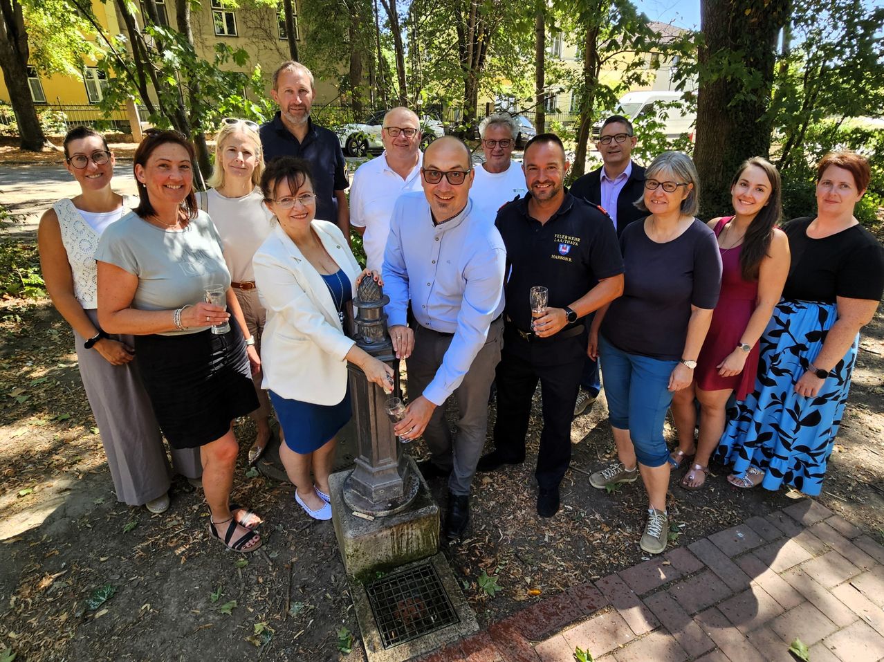 A group of people are gathered around a fountain in a park, smiling and holding glasses. They are dressed casually and appear to be celebrating an event.