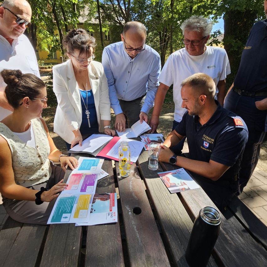 A group of people are gathered around a wooden table outdoors, reviewing documents. A man in a blue shirt is seated, while others stand and look at papers.