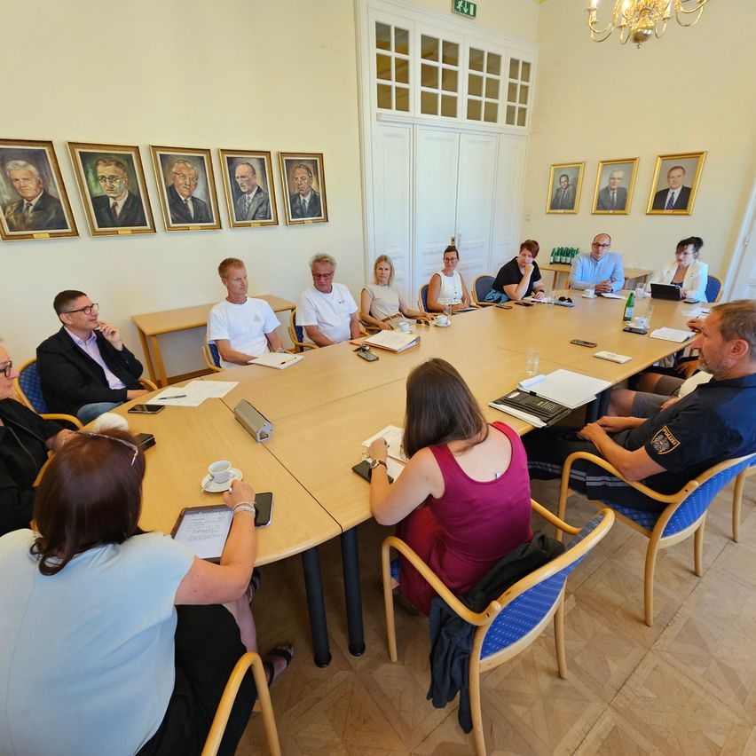 A group of people are sitting around a long table in a conference room, with several portraits of men on the wall behind them. They are engaged in a discussion, with some holding papers and others looking at laptops.
