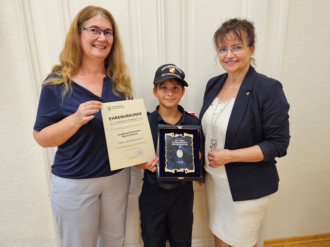Three individuals stand together, smiling for a photo. A boy in a uniform holds a framed award, while two women, one holding a certificate, flank him. The setting appears formal, possibly for an award ceremony.