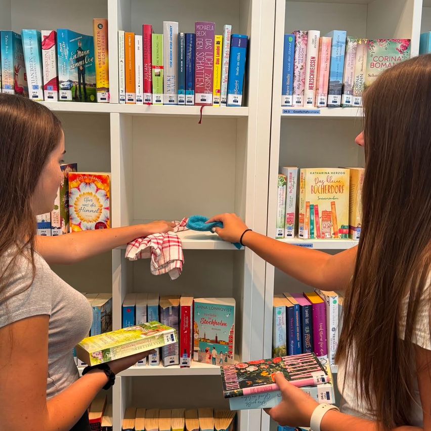Zwei Frauen reinigen ein Bücherregal in einer Bibliothek, das mit Büchern gefüllt ist. Eine Frau hält ein Buch und ein Tuch.