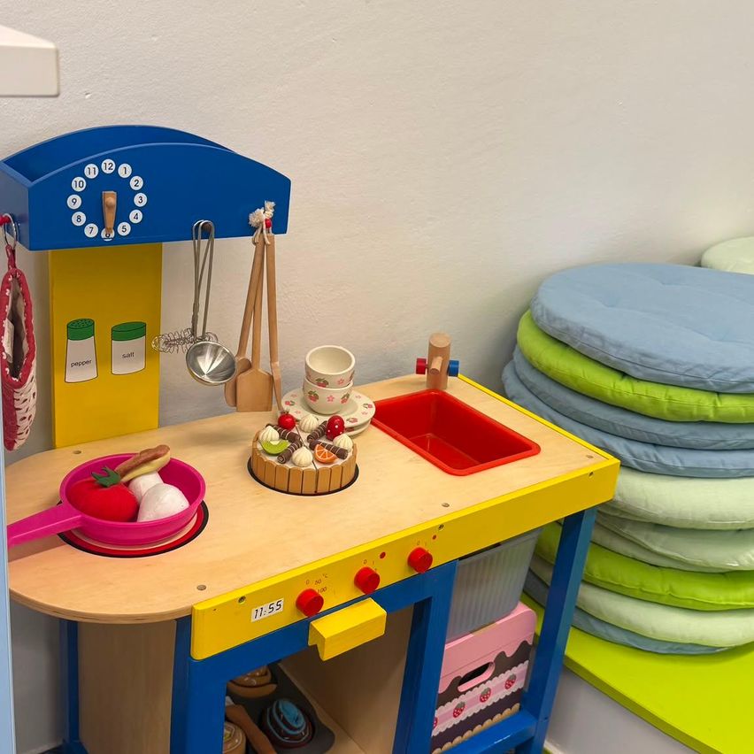 Eine farbenfrohe Spielküche mit Uhr, Utensilien und einem kleinen Kuchen auf einem Teller.