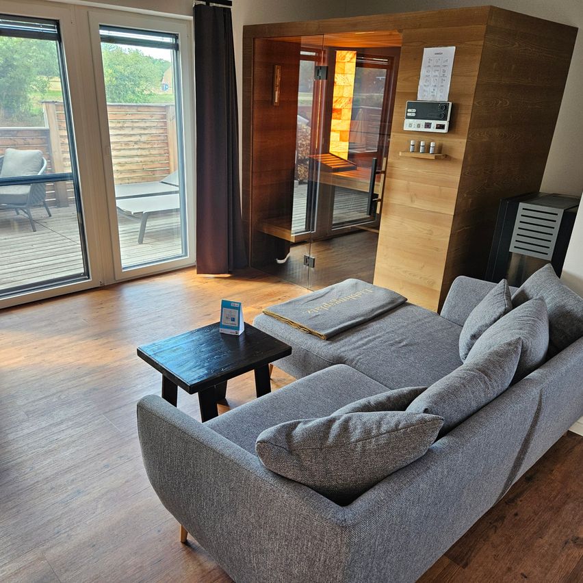 Ein modernes Wohnzimmer mit einem grauen Ecksofa, einem schwarzen Couchtisch und einer hölzernen Sauna im Hintergrund. Der Raum hat Holzböden und große Glastüren, die zu einer Terrasse führen.