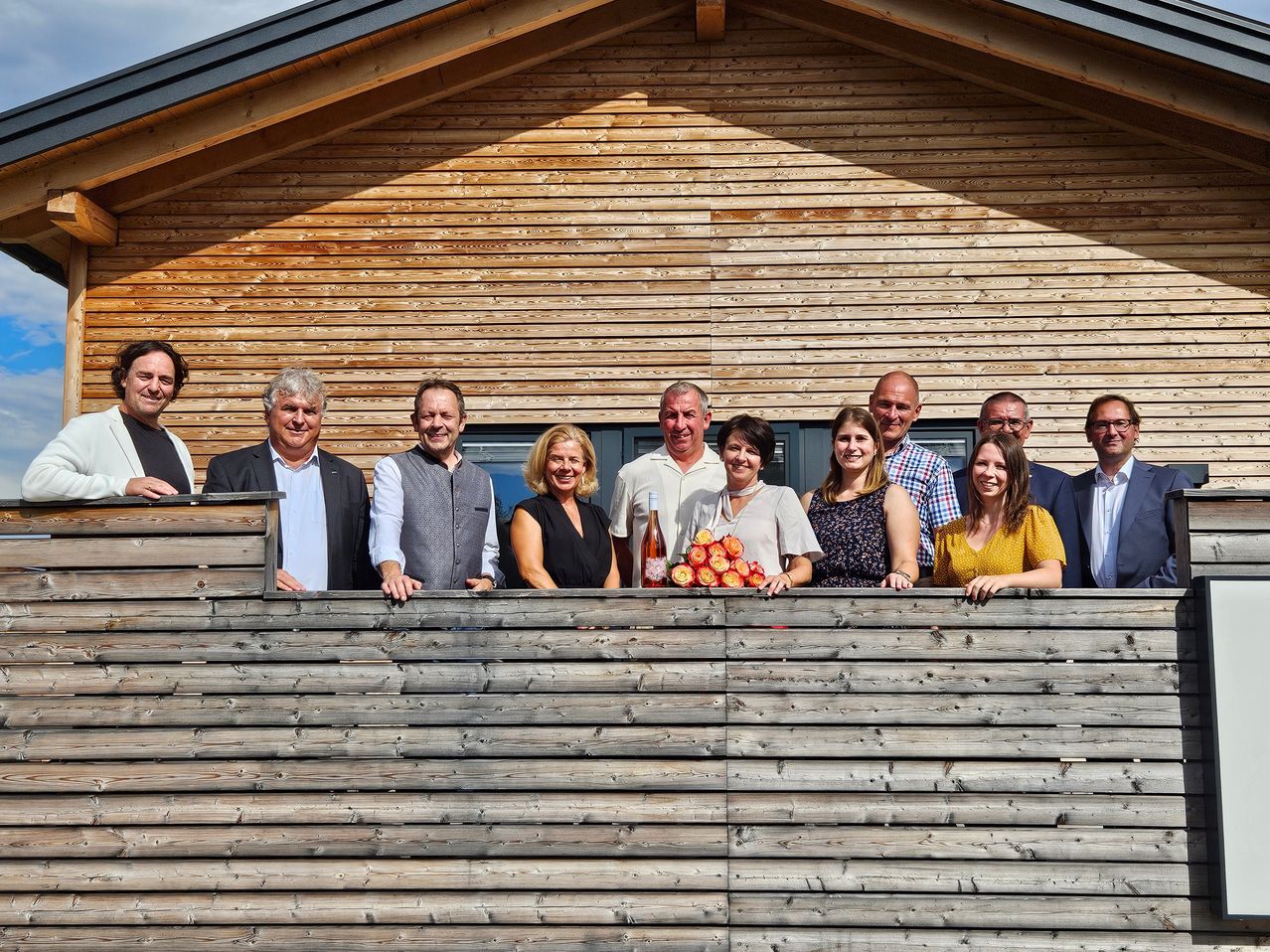 Eine Gruppe von Menschen, möglicherweise eine Familie oder ein Team, steht auf einem hölzernen Balkon vor einem Holzhaus. Sie lächeln und posieren für ein Foto.