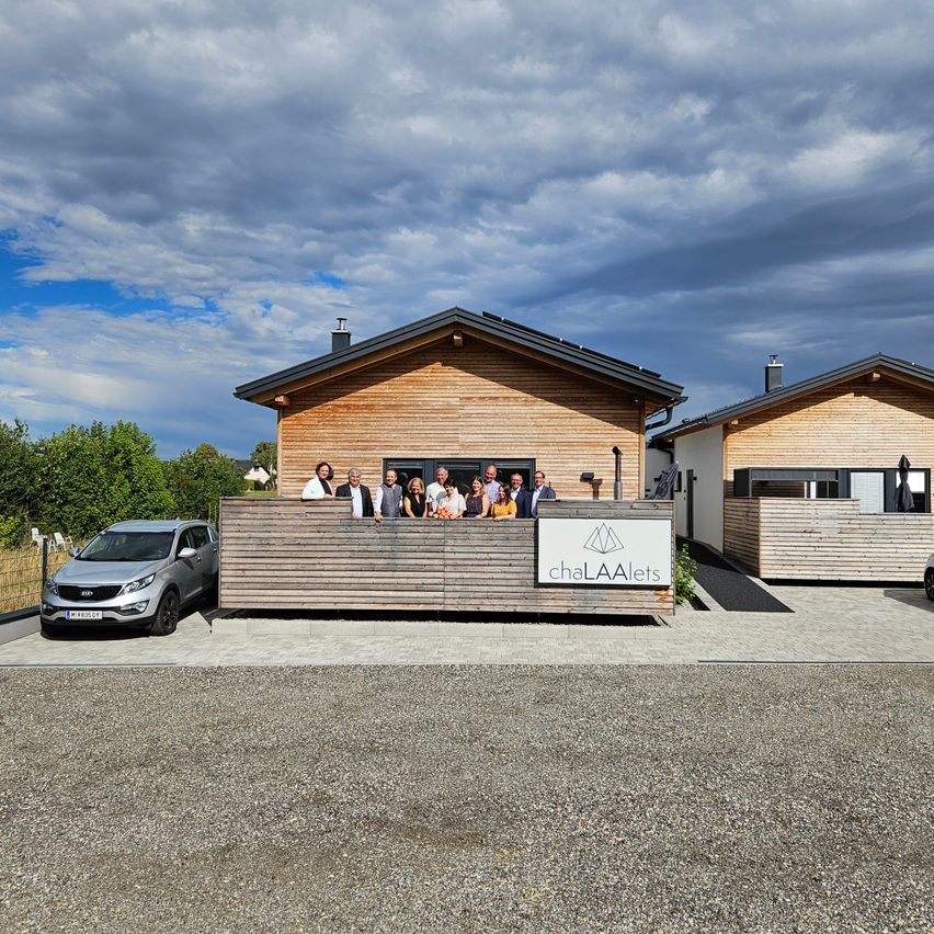 Eine Gruppe von Menschen posiert für ein Foto vor einem modernen Holzhaus mit dem Schild 'chaLAAlets'. Ein silberner Wagen ist in der Nähe geparkt.