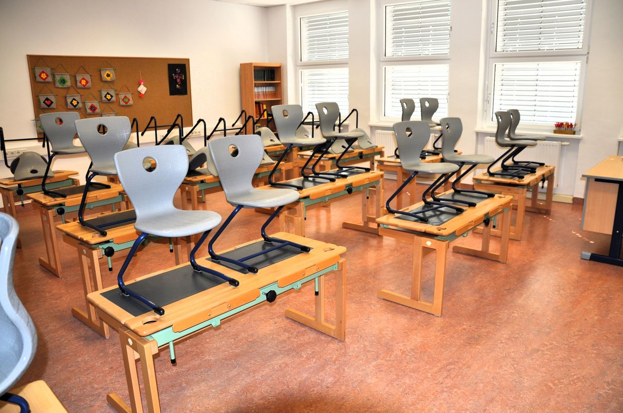 An empty classroom with gray desks and chairs arranged in rows, sunlight streaming through the windows.
