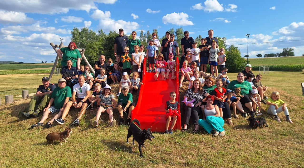A group of people, including adults and children, are gathered around a red slide in a grassy field. Some are sitting, some are standing, and some are holding balloons. The sky is blue with clouds.