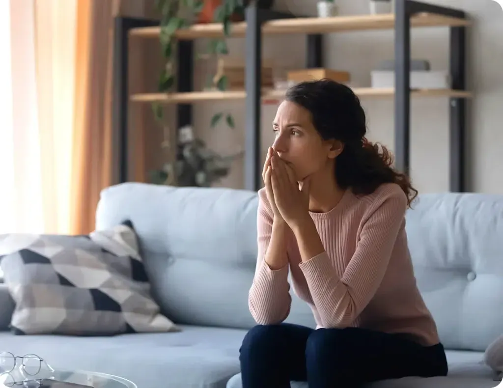 A woman in a pink sweater sits on a blue couch, her hands covering her mouth, looking thoughtful or worried. Behind her, a shelf with plants and a curtain is visible.