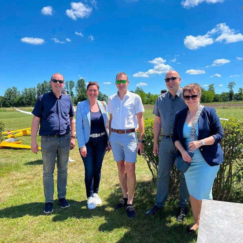 Fünf Menschen stehen an einem sonnigen Tag zusammen. Sie lächeln und scheinen für ein Foto zu posieren. Hinter ihnen sind Büsche und ein kleines Flugzeug auf dem Rasen.