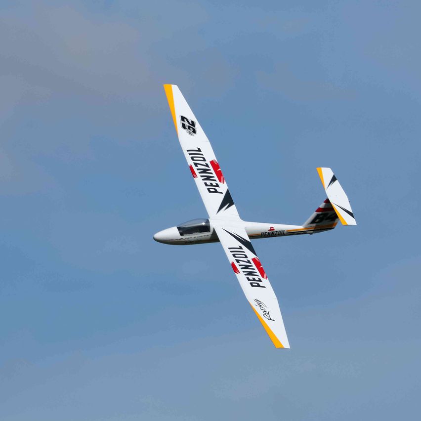 Ein ferngesteuertes Segelflugzeug mit 'PENNZOIL' und '25' Markierungen fliegt gegen einen blauen Himmel mit einigen Wolken.