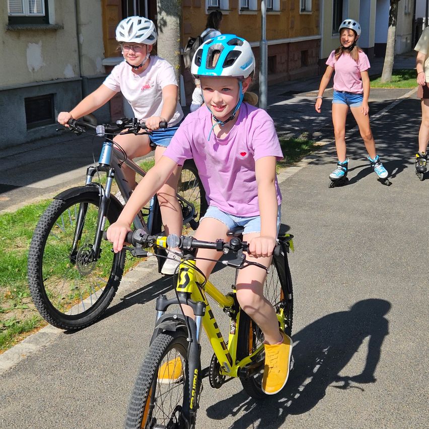 Drei junge Mädchen fahren auf einem asphaltierten Weg mit Fahrrädern und Rollschuhen. Das Mädchen vorne trägt einen Helm und ein lila Shirt. Dahinter fahren zwei weitere Mädchen mit Fahrrädern, und ein weiteres Mädchen fährt mit Rollschuhen.