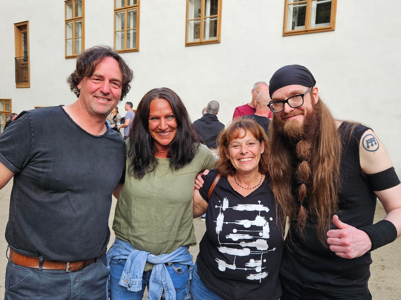 Vier Menschen stehen eng für ein Foto zusammen, mit zwei Frauen und zwei Männern. Der Mann auf der linken Seite trägt einen braunen Gürtel. Die Frau in der Mitte trägt ein grünes Hemd. Der Mann auf der rechten Seite hat lange Haare und einen Bart. Hinter ihnen steht ein Gebäude mit vielen Fenstern.