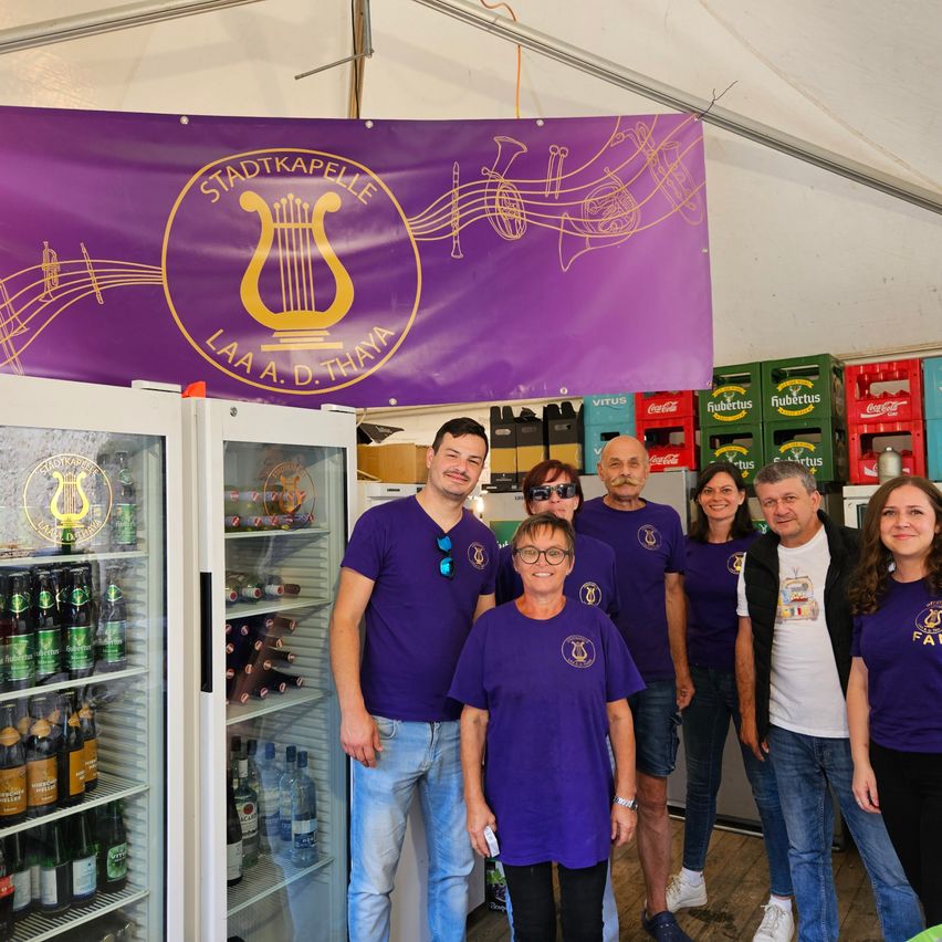 Eine Gruppe von Menschen in lila Shirts und mit Brillen steht hinter einem Kühler voller Getränke in einem Zelt mit einem lila Banner mit Musiknoten und einer Leier.