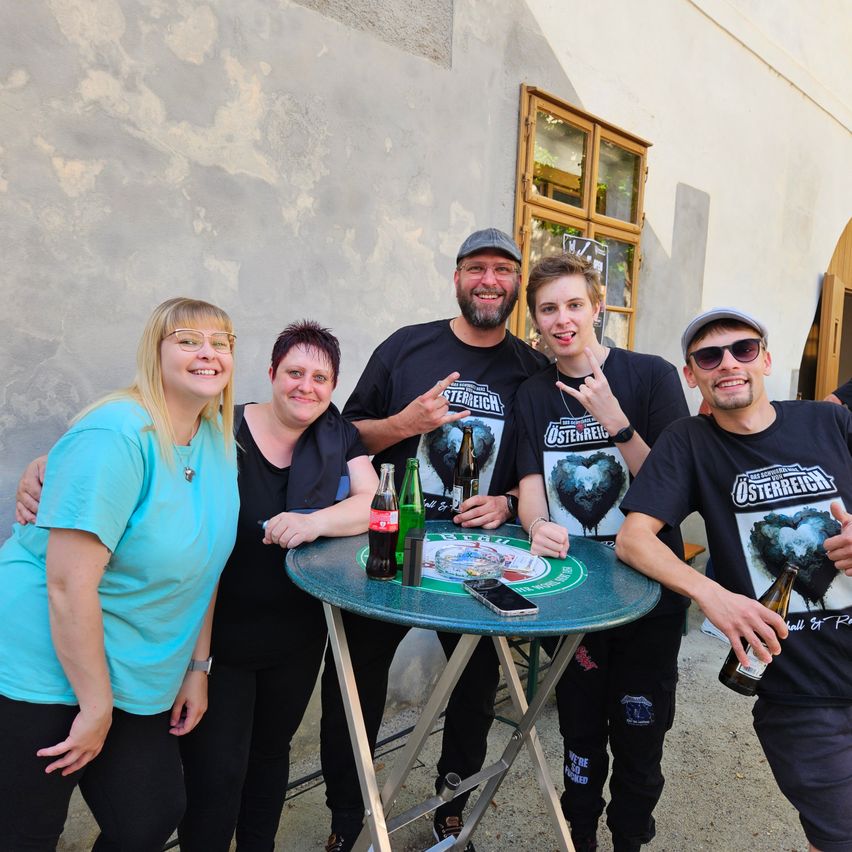 Eine Gruppe von Menschen posiert für ein Foto vor einem Gebäude mit Getränken und einem Tisch vor ihnen. Sie alle lächeln.