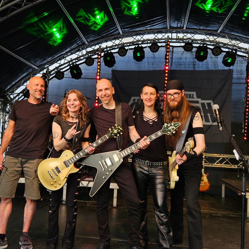 Eine Band steht auf einer Bühne mit grünen Lichtern darüber. Die Bandmitglieder halten Gitarren und lächeln für das Foto. Dahinter ist ein Hintergrund mit einem Logo.