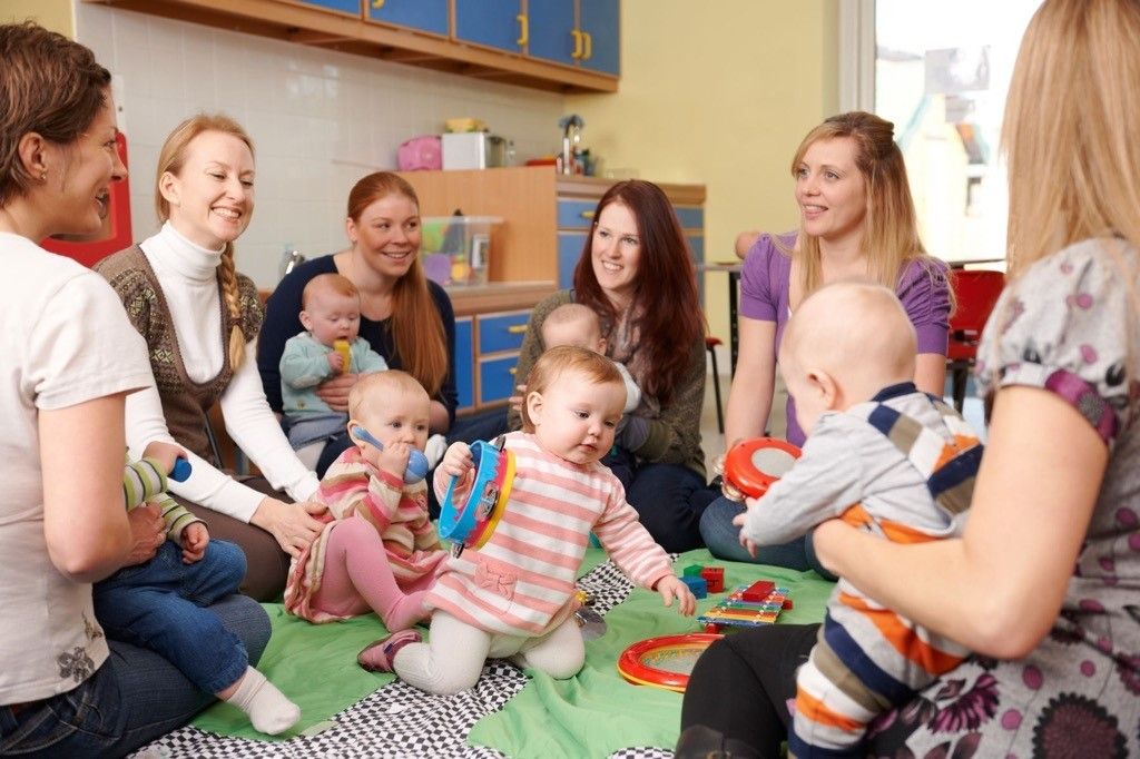 Fünf Frauen und fünf Babys sind in einem Raum versammelt. Die Frauen lächeln und interagieren mit den Babys. Spielzeug liegt auf dem Boden und der Raum hat Schränke und ein Fenster.