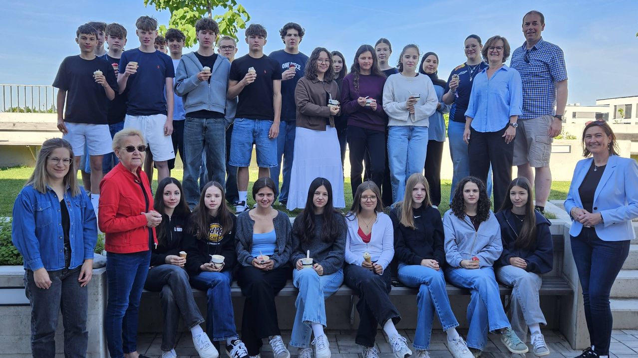 Eine Gruppe von Schülern, sowohl Jungen als auch Mädchen, lässig gekleidet, einige mit Brille, posieren für ein Foto mit Eiscreme, während sie auf einer Bank und Gras sitzen und stehen.