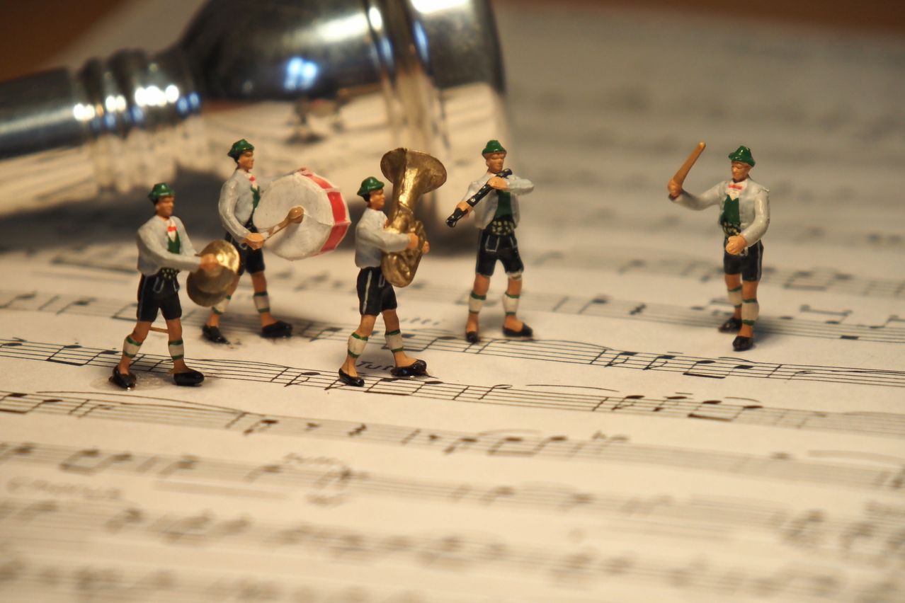 Fünf Spielzeugmusiker, in grünen Hüten und Shorts, spielen Trommeln, eine Trompete und ein Tenorhorn auf einem Musikblatt. Ein silberner Shaker ist links oben.