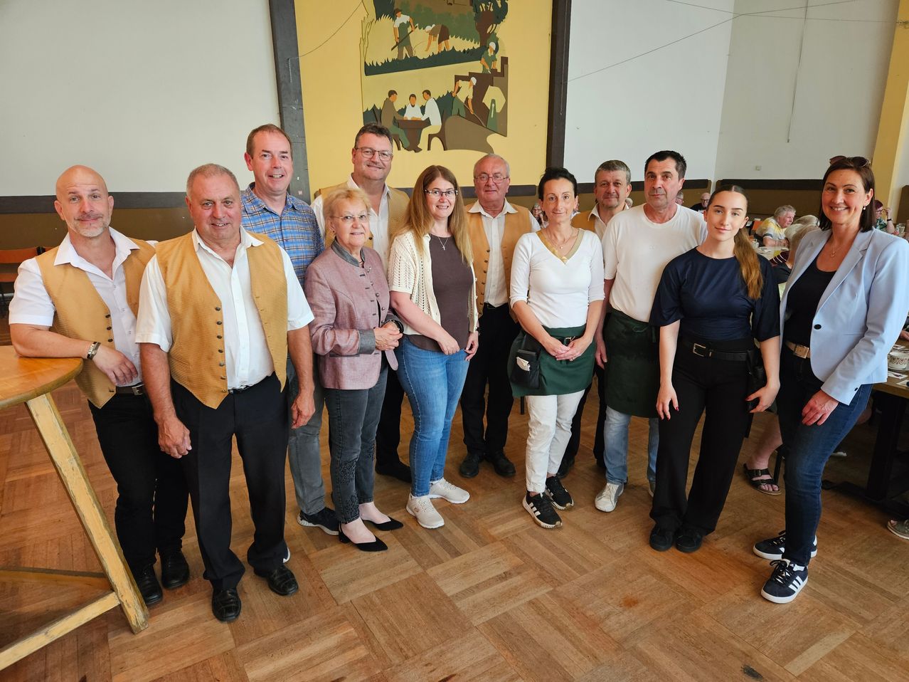 Eine Gruppe von Menschen, sowohl Männer als auch Frauen, posiert für ein Foto in einem Raum mit Holzboden. Einige tragen Brillen und Halsketten.