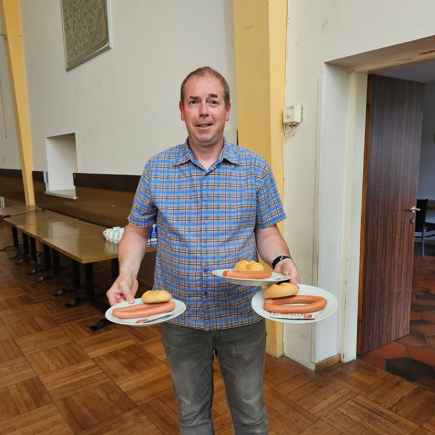 Ein Mann steht in einem Raum, lächelt und hält zwei Teller mit Essen. Ein Teller hat zwei Hot Dogs und der andere ein Sandwich. Hinter ihm stehen Holzbänke und eine Holztür.