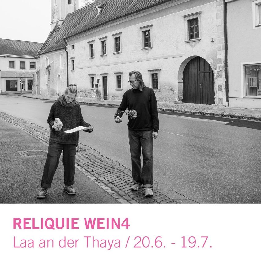 Zwei Personen stehen auf einer Straße mit alten Gebäuden im Hintergrund. Einer hält Papier, der andere einen Ball. Das Bild enthält Text für eine Ausstellung: 'Relique Wein4 Laa an der Thaya'.