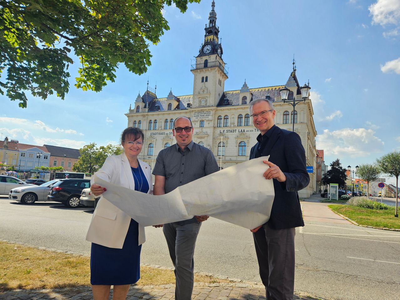 Drei Personen stehen vor einem großen Gebäude und halten ein großes weißes Papier in der Hand. Sie lächeln für die Kamera.