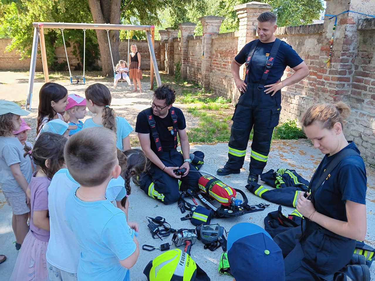 Zwei Feuerwehrleute demonstrieren Ausrüstung für eine Gruppe von Kindern in einem Park. Ein Feuerwehrmann kniet, während der andere hinter ihm steht. In der Nähe beobachtet eine Frau ein Kind auf einer Schaukel.
