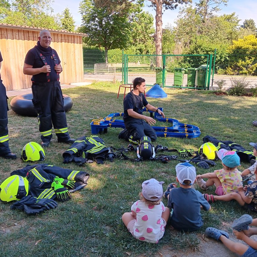 Zwei Feuerwehrleute geben Kindern auf einem Rasenfeld eine Lektion. Sie tragen Uniformen, Helme und Handschuhe. Die Kinder sitzen auf dem Rasen und lauschen.