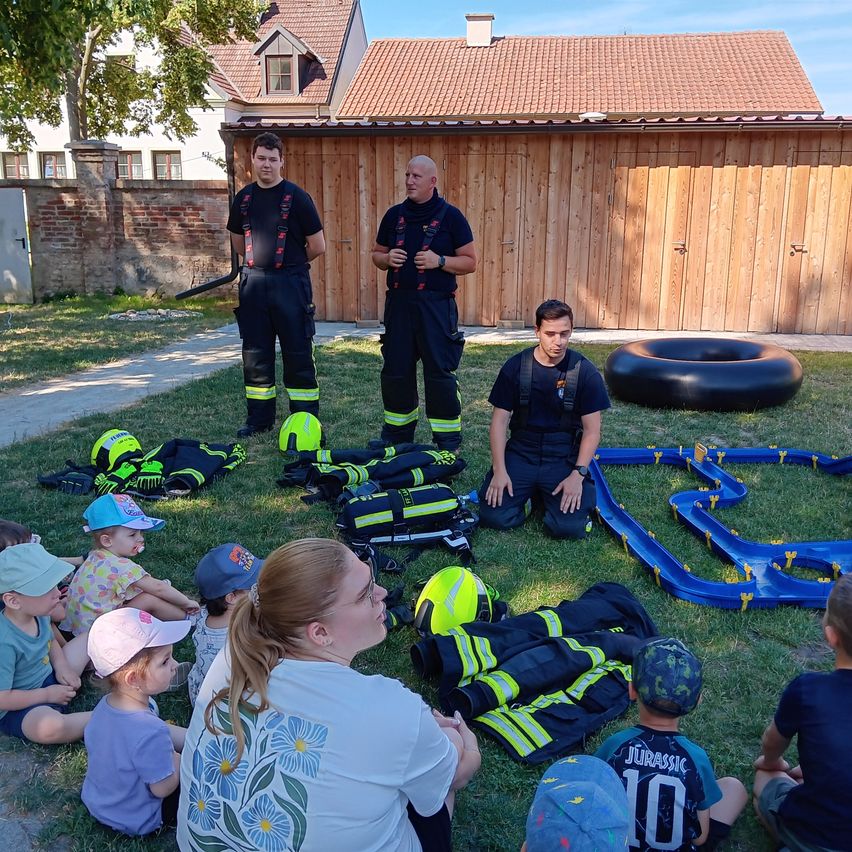 Feuerwehrleute demonstrieren Kindern auf einem Rasen mit einer Reifenspur, Helmen und Feuerlöschern die Brandschutzmaßnahmen.
