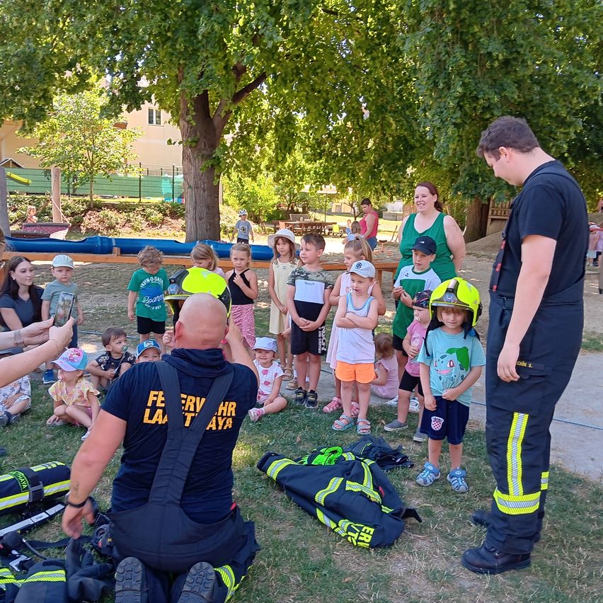 Feuerwehrleute unterrichten Kinder in einem Park mit Bäumen, einer Rutsche und einem Zaun. Einige Kinder tragen Mützen und Turnschuhe.
