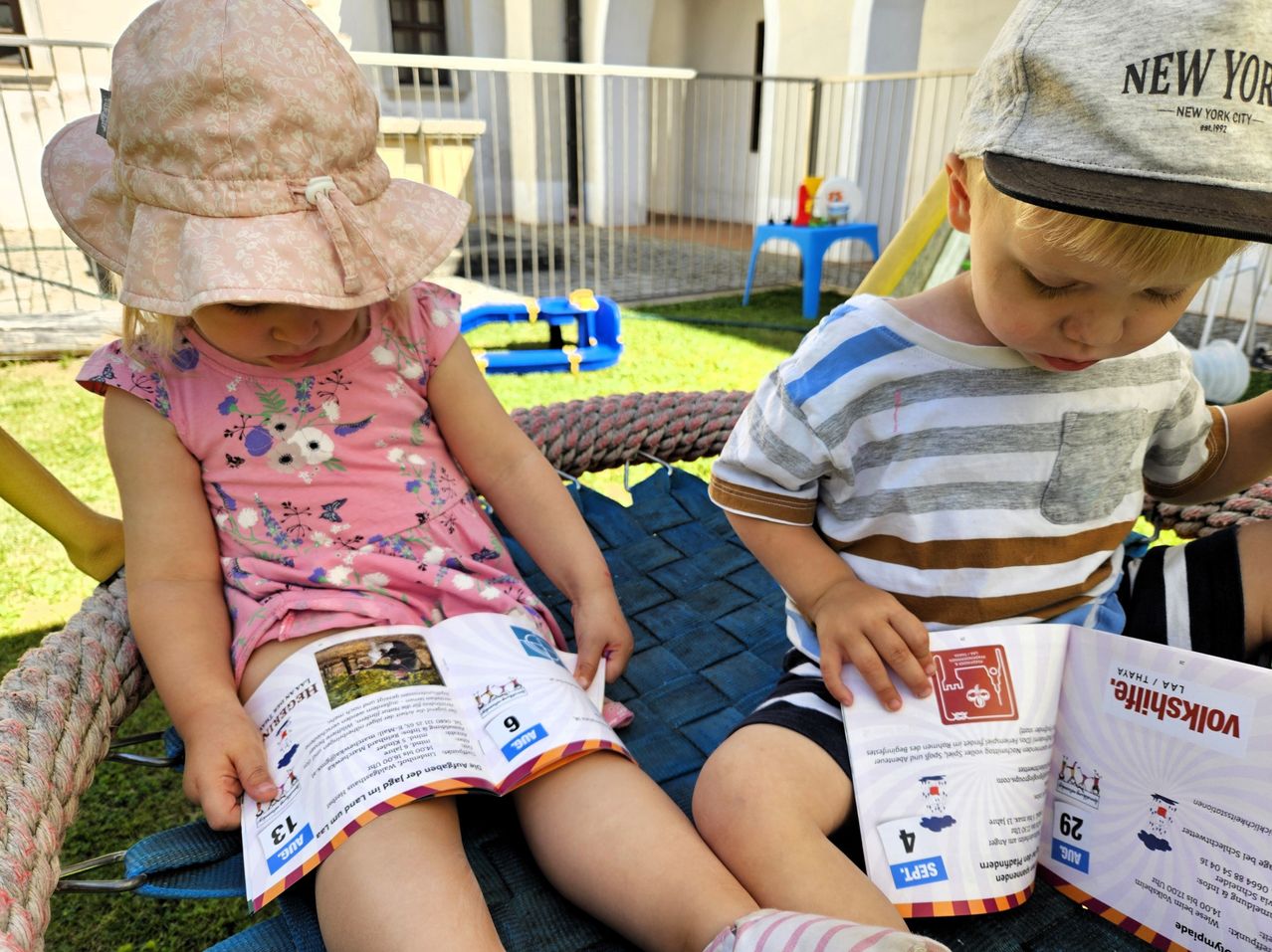 Zwei Kinder sitzen auf einer Matte im Garten. Ein Mädchen hält ein offenes Buch und ein Junge hält eine Broschüre. Sie scheinen zu lesen. Hinter ihnen sind Spielzeuge und ein Zaun.
