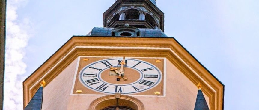 Ein hoher Glockenturm mit einer goldenen Uhr mit römischen Ziffern unter einem klaren blauen Himmel.