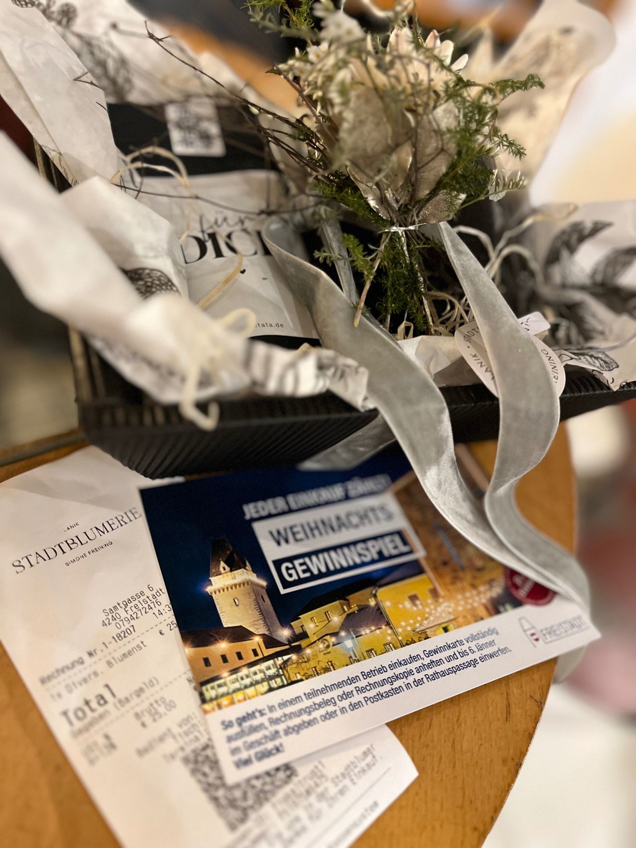 Ein kleiner Korb enthält einen Pflanzenstrauß und eine Schleife. Ein Ticket für Stadtblumerei und ein Weihnachtsgewinnspiel-Flyer liegen auf einer Holzoberfläche.