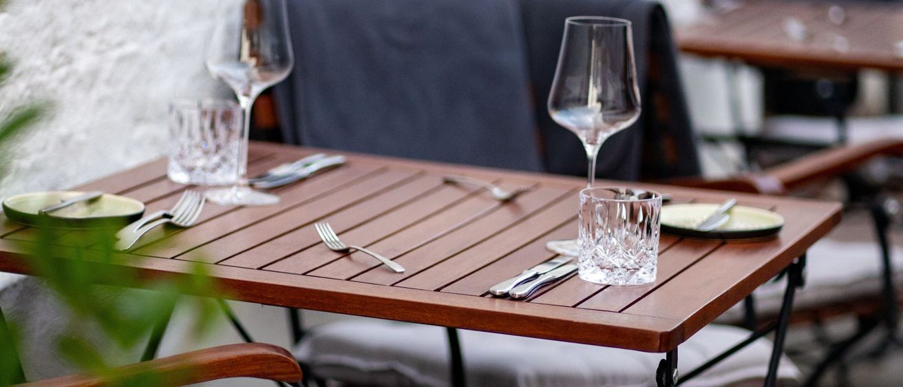 Ein Outdoor-Restauranttisch mit Weingläsern, Tellern und Besteck. Der Tisch ist aus Holz mit Metallbeinen und die Stühle sind gepolstert mit blauen Bezügen.