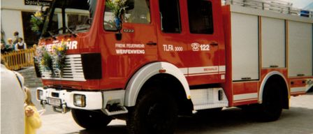 Ein roter Feuerwehrwagen mit der Nummer 1222 steht vor einem Gebäude. Er hat einen Blumenstrauß auf der Frontscheibe.