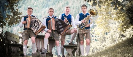Vier Männer in traditioneller Kleidung, mit Musikinstrumenten, posieren für ein Foto unter einem Baum, mit einer Holzbank und einem Berg im Hintergrund.