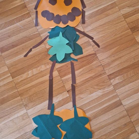 Ein Papierausschnitt eines pumkinförmigen Wesens mit braunen Stielen und Blättern liegt auf einem Holzboden. Die Figur hat ein Gesicht aus braunem Papier.