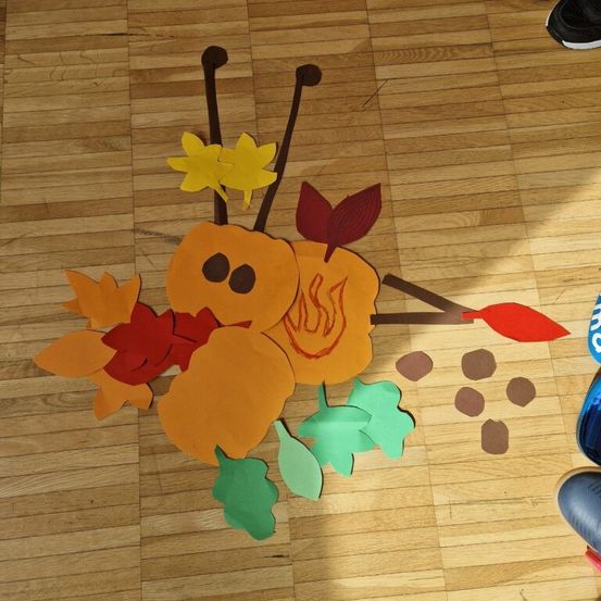 Eine handgefertigte Herbstdekoration aus Papierblättern und Kürbissen liegt auf einem Holzboden. Die Farben umfassen gelbe, rote und orange Blätter.