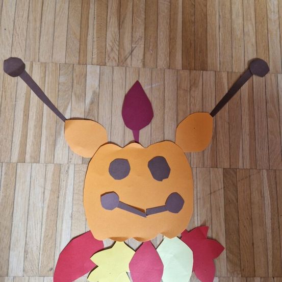Ein Papierausschnitt einer Figur mit Kürbiskopf, roten Ohren und einem roten Blatt auf einem Holzboden.