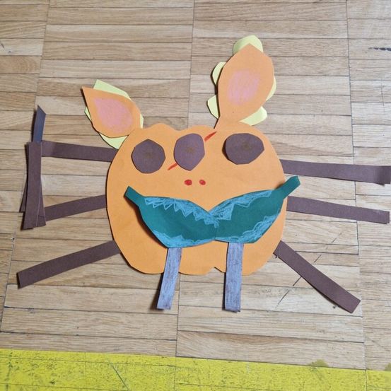 Ein Papierbastelwerk, das einem Kürbis mit einem grünen Mund ähnelt, auf einem Holzboden, aus braunem Papier hergestellt.