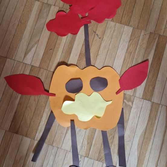 Eine Papierkürbis-Bastelarbeit mit roten Blättern und einem gelben Lächeln, auf einem Holzboden platziert.