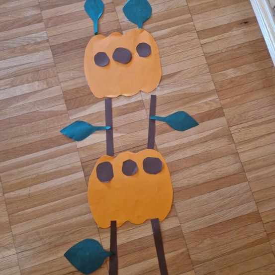 Zwei Papierkürbisse mit braunen Stielen und Blättern sind auf einem Holzboden angeordnet. Jeder Kürbis hat ein Gesicht mit zwei Augen und einem Mund.