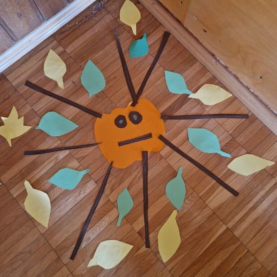 Ein orangefarbener Kürbiskopf wird auf einem Holzboden mit Papierblättern und Stöcken gestaltet. Der Kürbis hat einen traurigen Ausdruck, umgeben von bunten Blättern.