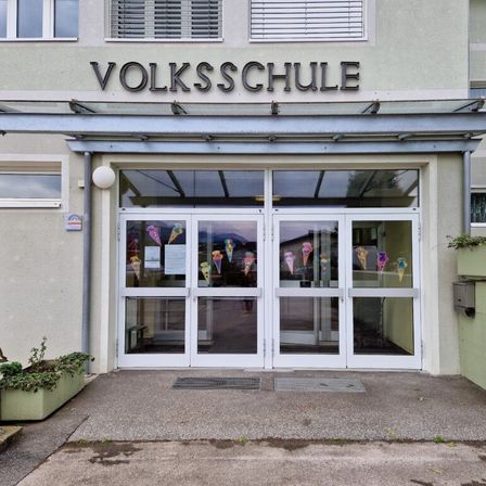 Der vordere Eingang eines Gebäudes mit der Aufschrift 'Volksschule' mit Glastüren und Fenstern, Eis-Themen-Dekorationen und einem kleinen Schild an der Wand.