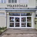 Der vordere Eingang eines Gebäudes mit der Aufschrift 'Volksschule' mit Glastüren und Fenstern, Eis-Themen-Dekorationen und einem kleinen Schild an der Wand.