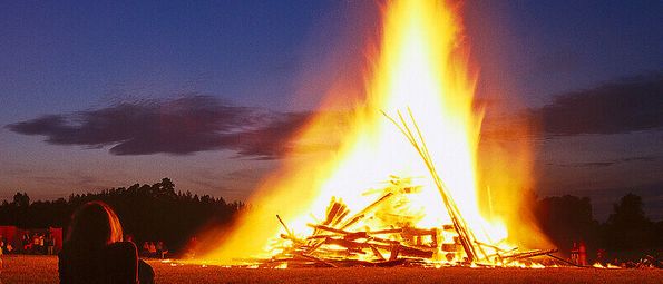 Ein großes Lagerfeuer brennt hell in der Nacht, umgeben von Zuschauern auf einer grasbewachsenen Wiese unter einem blauen Himmel mit Wolken.