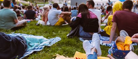 Eine große Gruppe von Menschen sitzt auf einem Grasfeld und schaut sich einen Film auf einer großen Leinwand an. Sie sind lässig gekleidet und haben Decken und Taschen um sich herum.