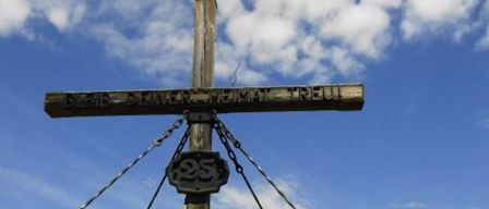 Ein hölzerne Kreuz steht hoch mit einer Metallplatte mit der Nummer 25, verbunden durch Ketten, vor dem Hintergrund eines blauen Himmels mit verstreuten Wolken.
