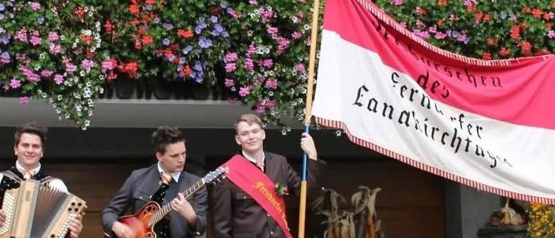 Vier Männer in Anzügen spielen Instrumente und halten eine Fahne auf einer Bühne, wahrscheinlich für ein traditionelles Ereignis. Die Fahne steht für 'Landwirtschaftstag'.